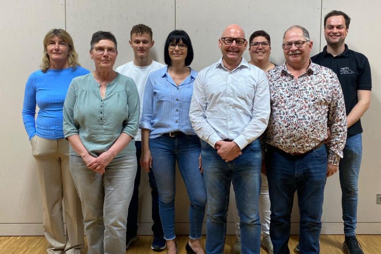 Die Vorstandschaft des Musikvereins. Gruppe von neun Personen, lächelnd, vor einer neutralen Wand.
