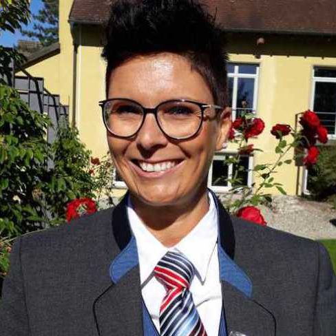 2. Vorständin. Frau mit schwarzen kurzen Haaren, lächelnd in  Uniform des Musikverein Rheinheim. Gelbes Haus im Hintergrund und rote Rosen