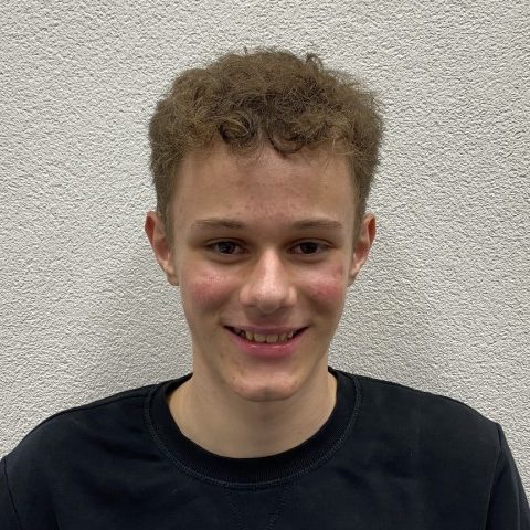 Theo Sutter, Beisitzer. Junger Mann mit lockigem Haar, lächelt vor einer grauen Wand.