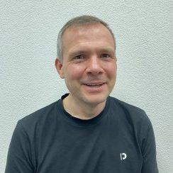 Dirigent Sascha Noll. Mann mit kurzem, grauem Haar lächelt vor einer hellen Wand. Trägt ein schwarzes T-Shirt.